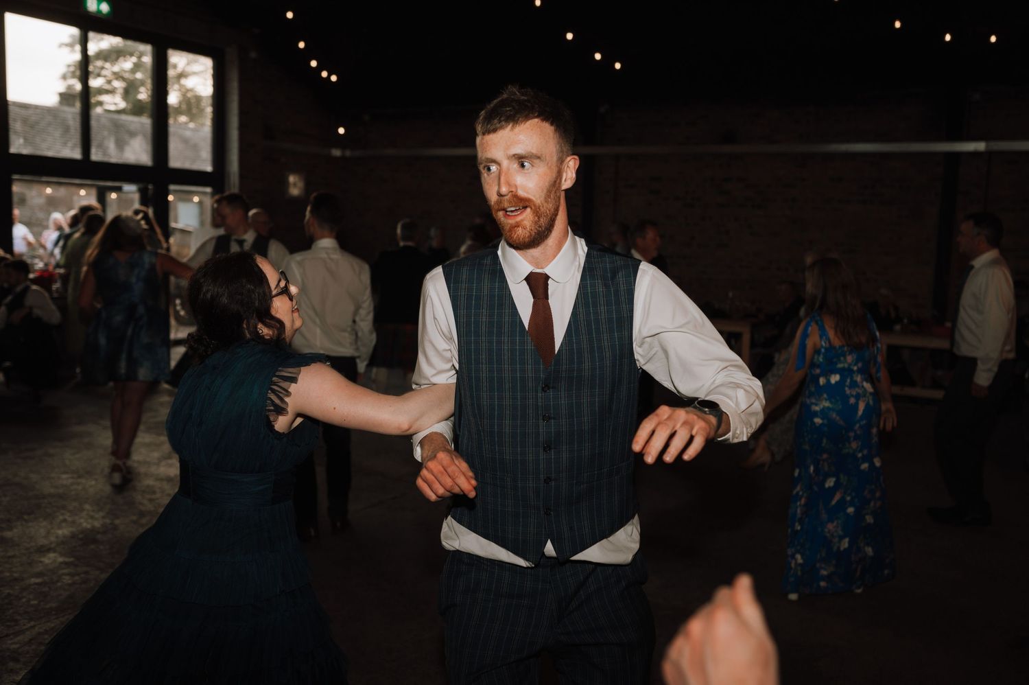 A guest in formal attire dances at a Scottish wedding reception in a rustic brick venue.
