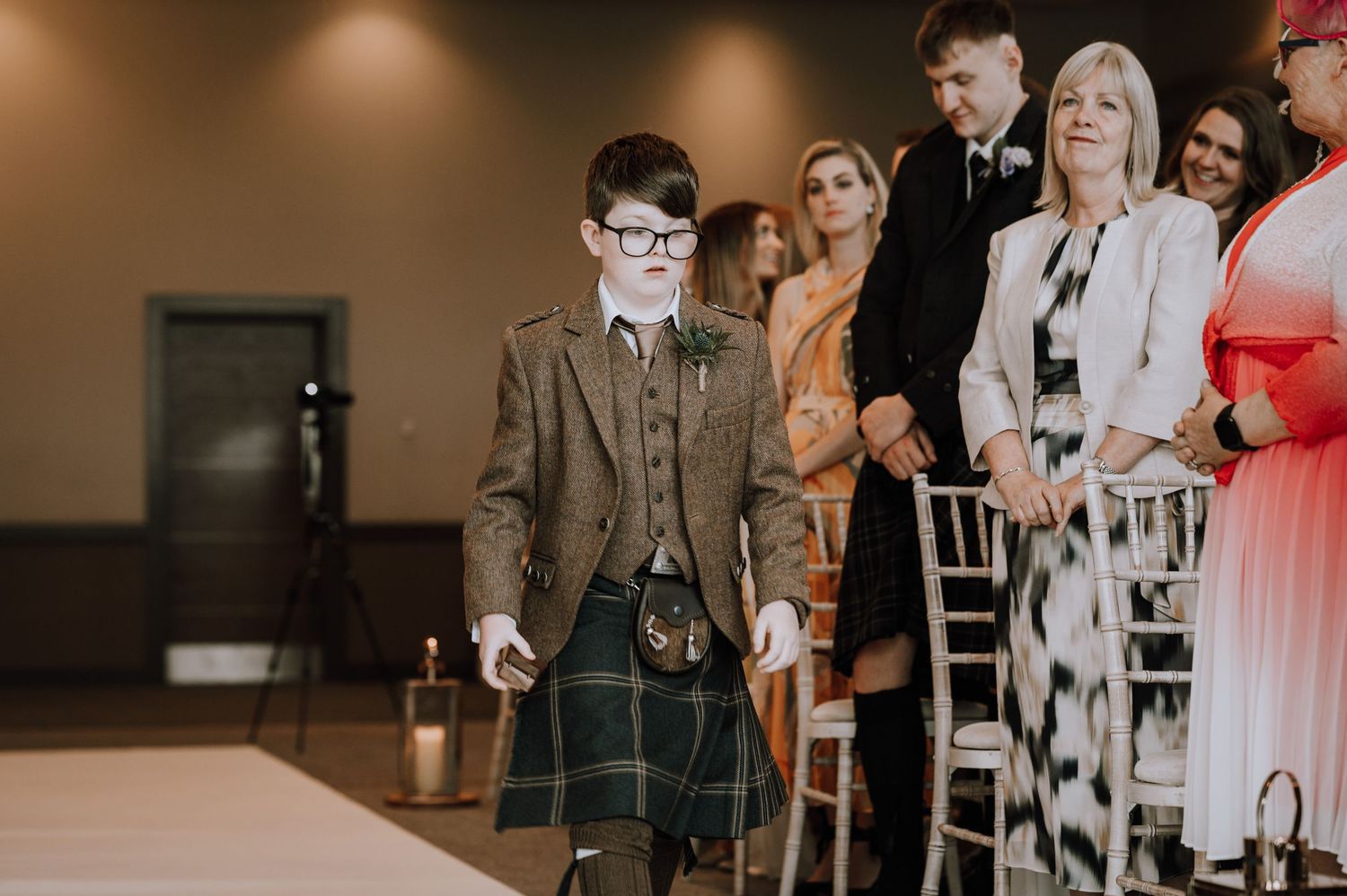 A young attendee in a traditional Scottish kilt and suit walks down the aisle at a formal wedding ceremony.