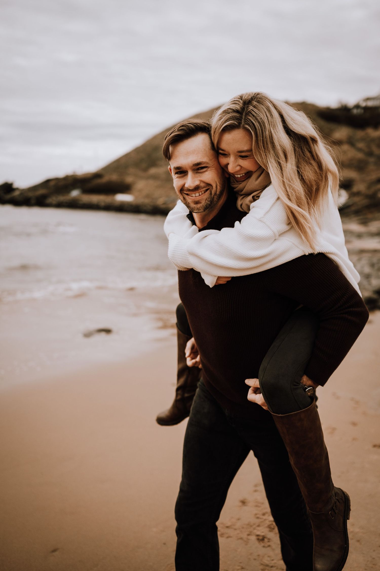 A playful piggyback ride moment captured on a misty beach with cliffs in the background.