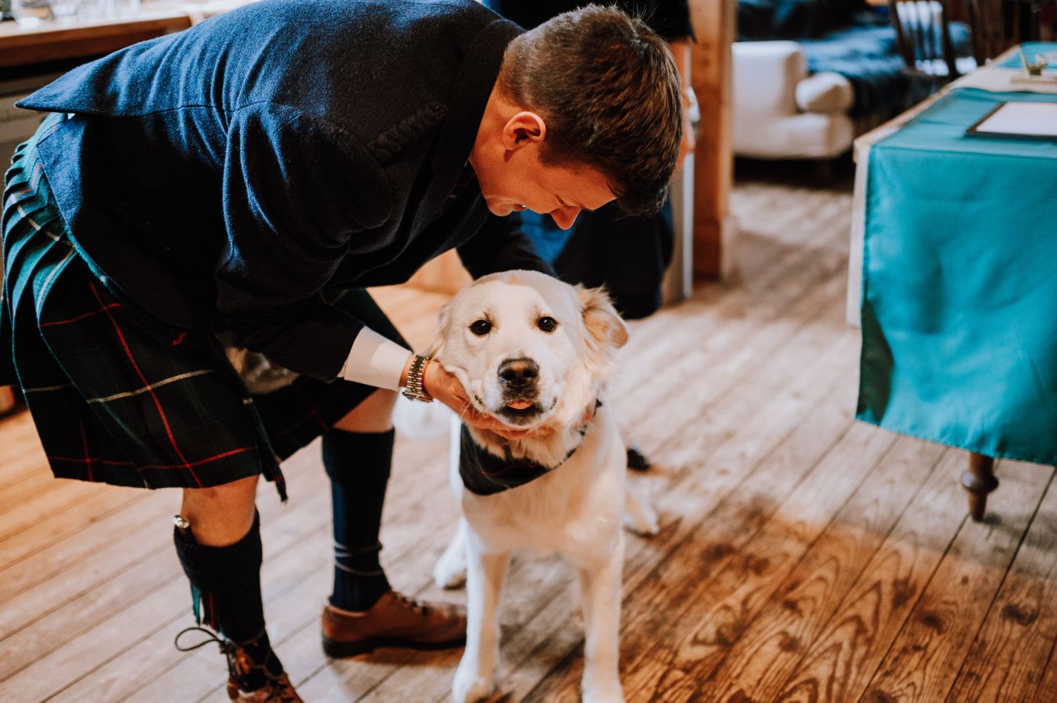 Someone in traditional Scottish attire pets a white dog wearing a bowtie on wooden floors.