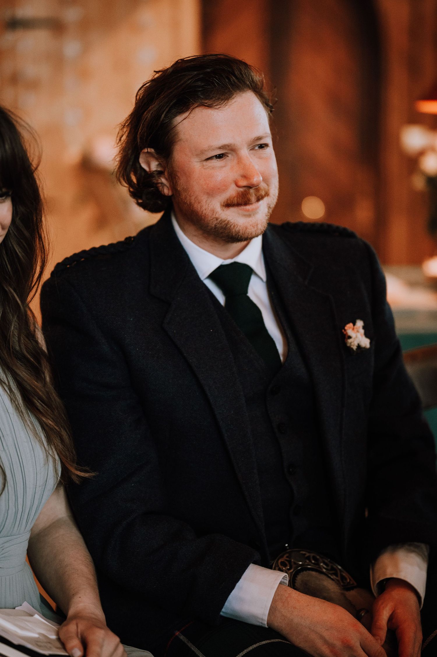 A guest at a wedding smiles warmly while wearing a black suit and tie.