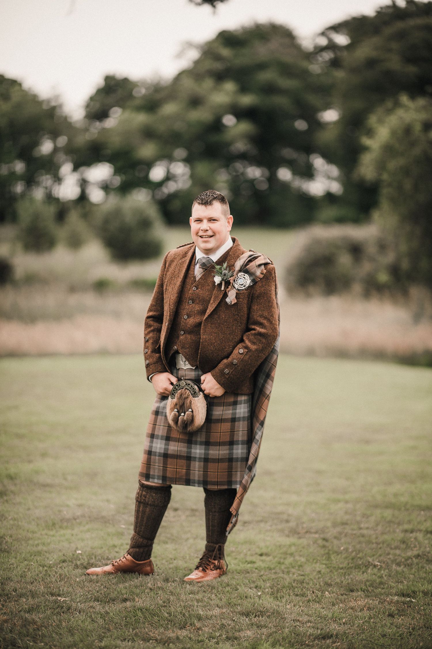 Man in brown tweed jacket and traditional Scottish kilt stands on grass at outdoor event.