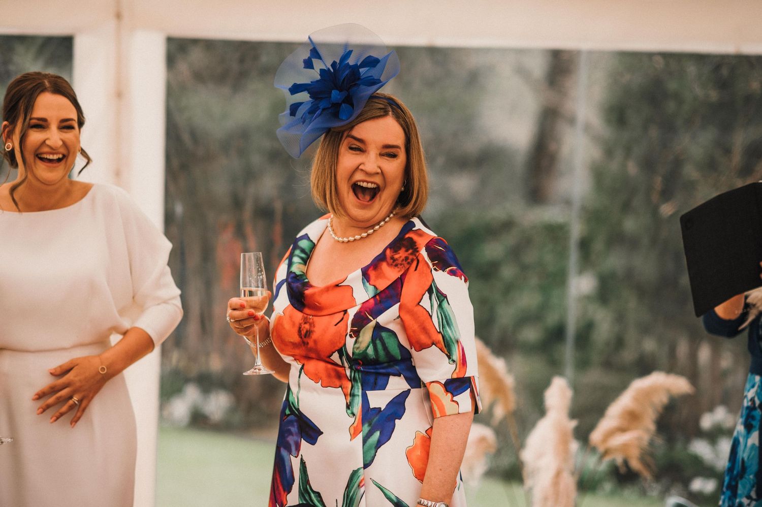 Two wedding guests share a celebratory moment outdoors, with one wearing a vibrant floral dress and blue fascinator.