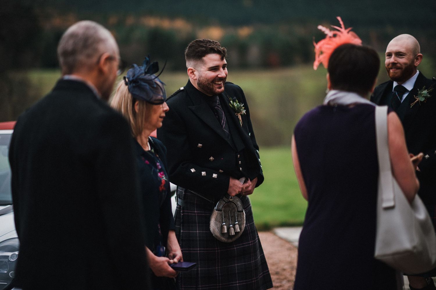 Wedding guests in kilts and formal wear gather for conversation outdoors.