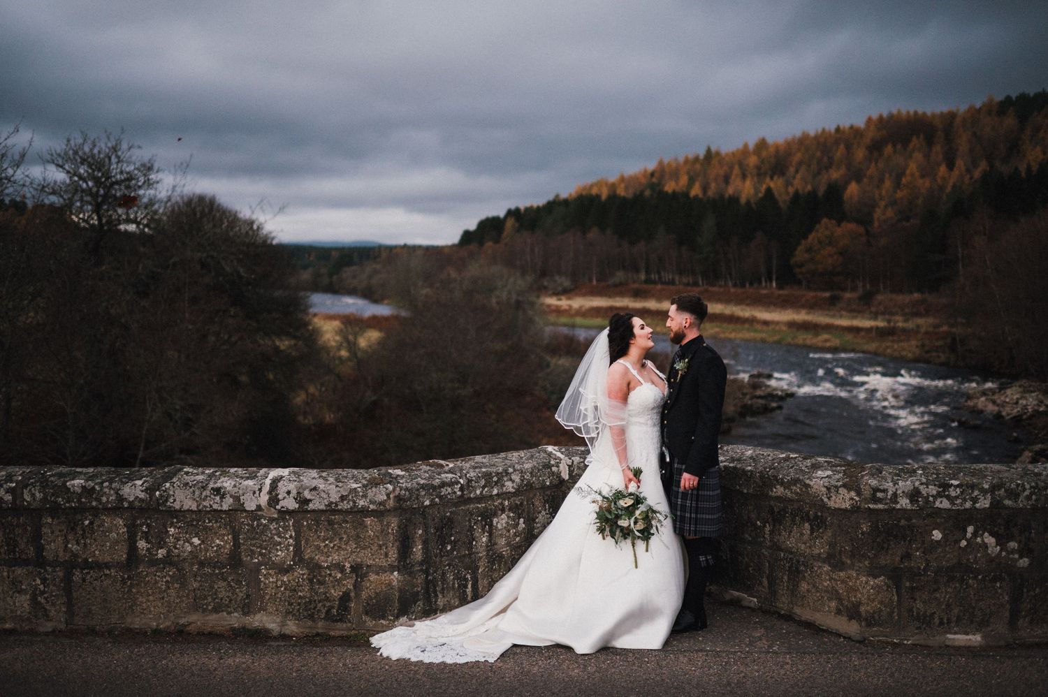 Wedding couple embraces on historic stone bridge over flowing river.