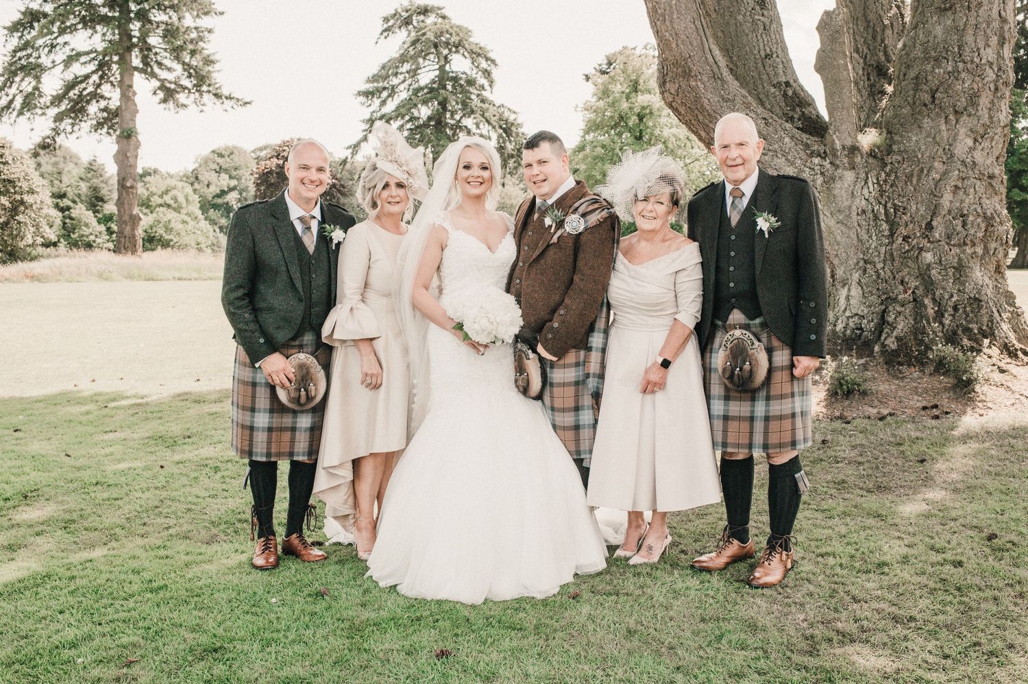 Wedding party in traditional Scottish attire posing under large trees.