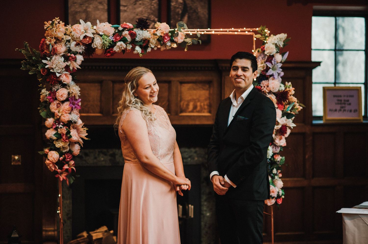 A ceremony scene under a floral arch with pink and white blooms against dark wood paneling.