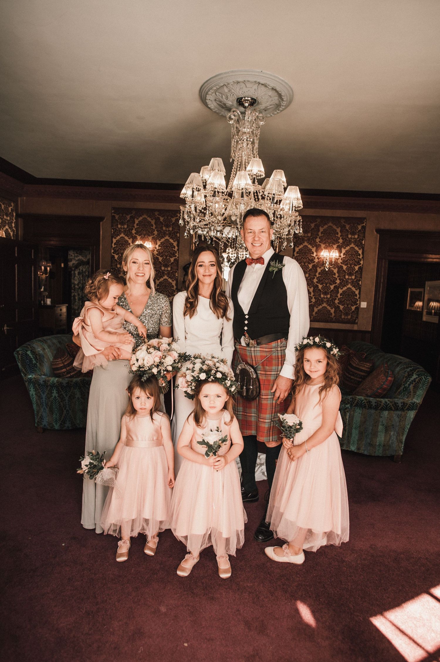 Wedding party poses under crystal chandelier in elegant room with traditional Scottish attire.