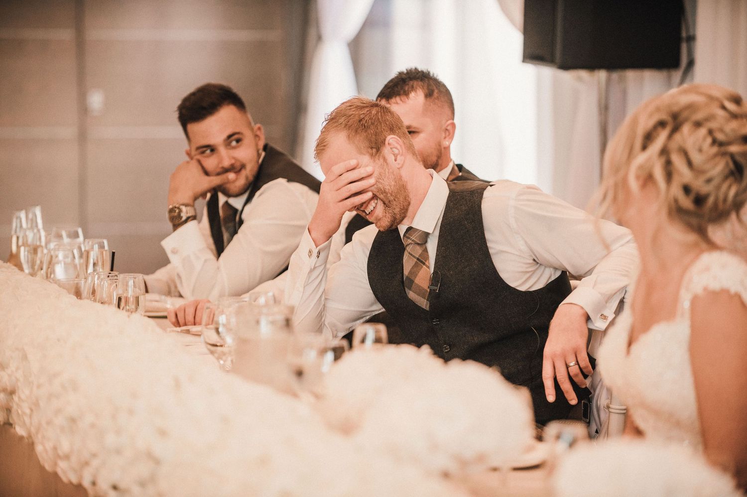 Wedding party members react humorously during reception speeches at an elegant white-decorated venue.
