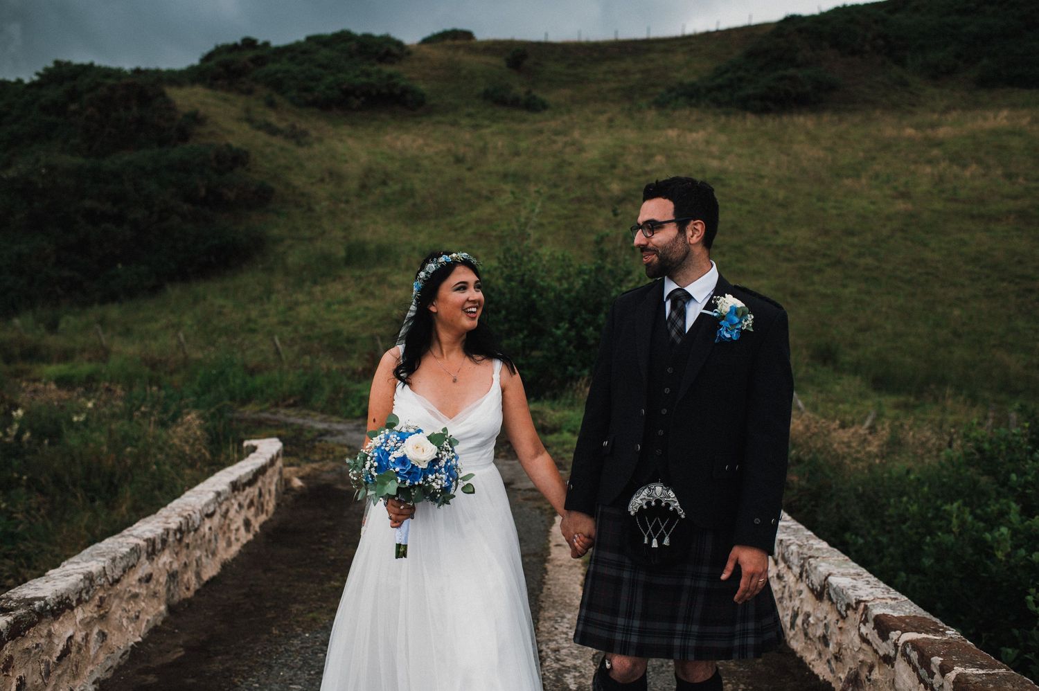 A couple in traditional Scottish wedding attire pose together on a rustic stone bridge.
