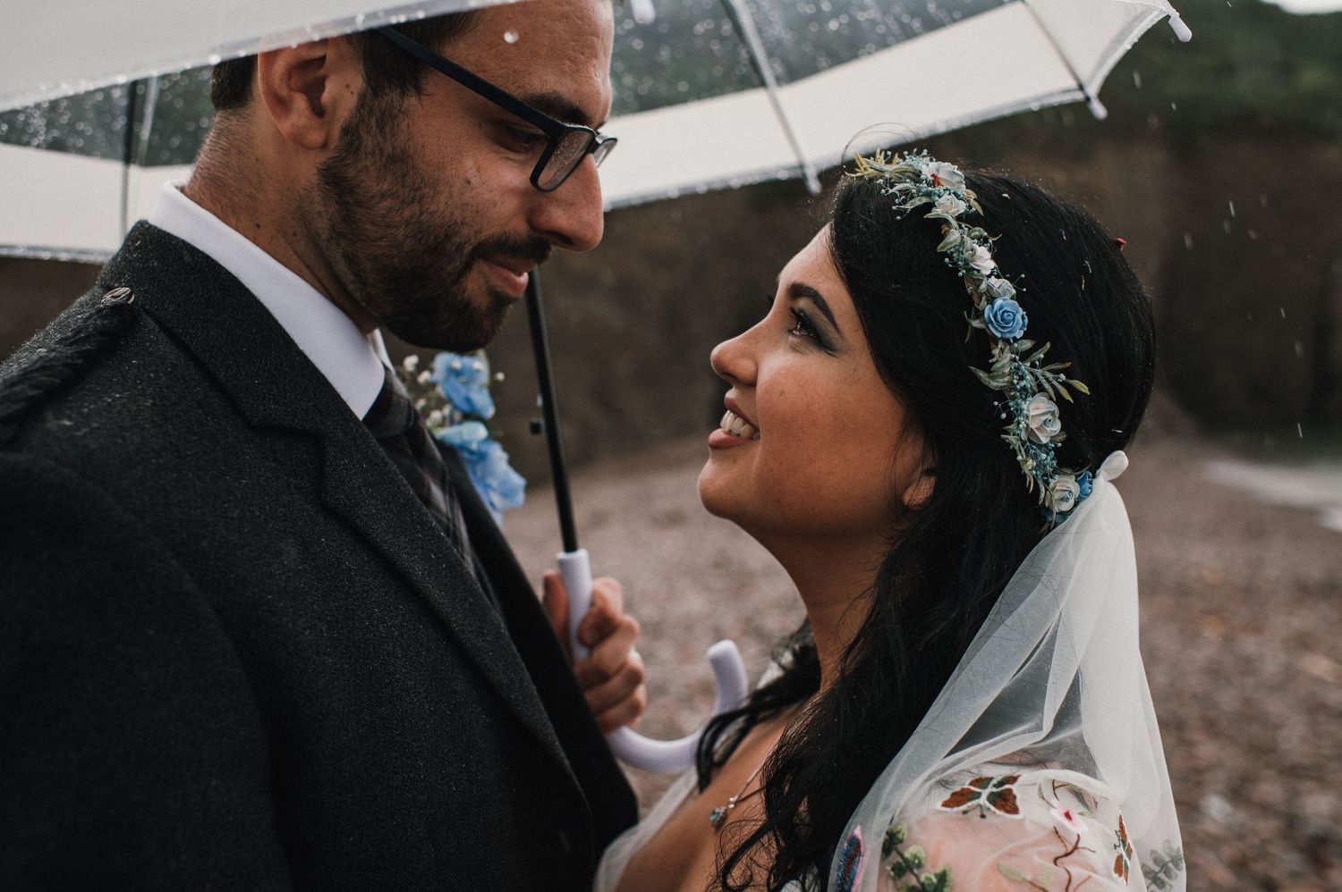 A couple shares an intimate moment under a clear umbrella on their rainy wedding day.
