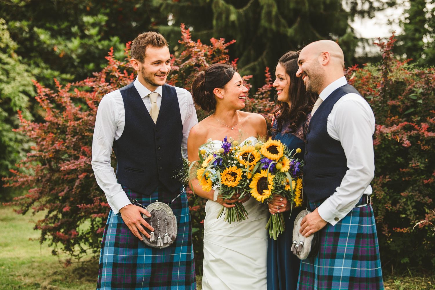 Wedding party in Scottish kilts and formal wear gather around sunflower bouquet outdoors.