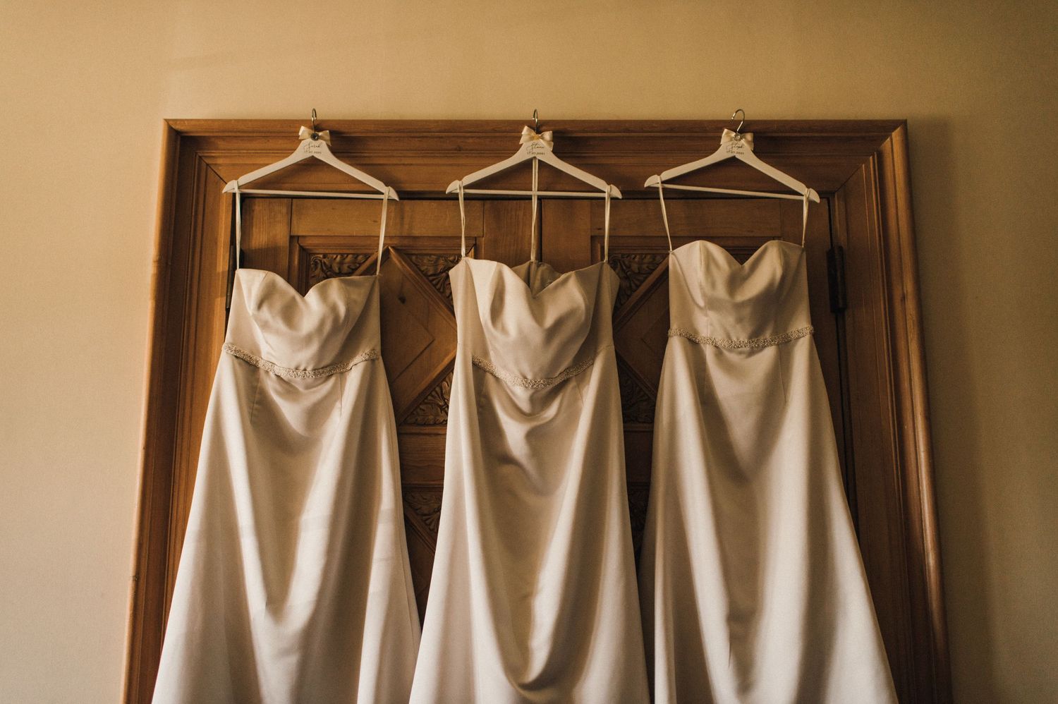 Three white bridesmaid dresses hanging on wooden hangers against brown wall.
