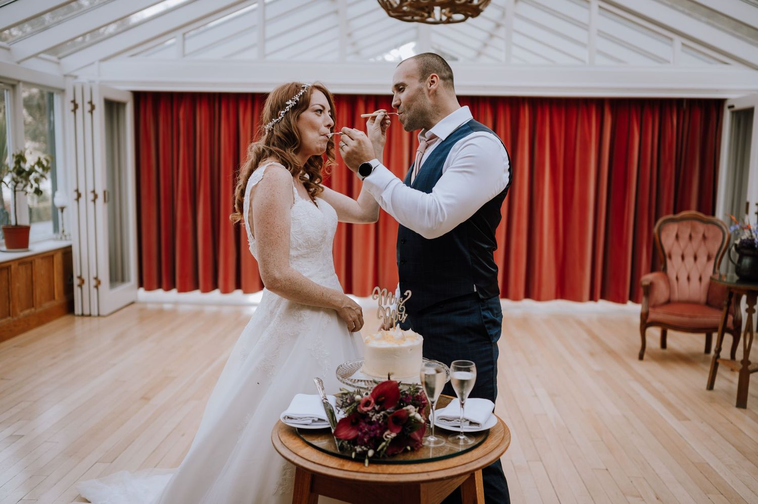 Newlyweds share cake during an intimate reception in a bright conservatory space.