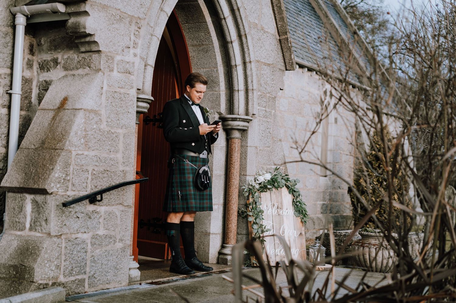 Person wearing traditional Scottish kilt and formal attire standing in stone church doorway.