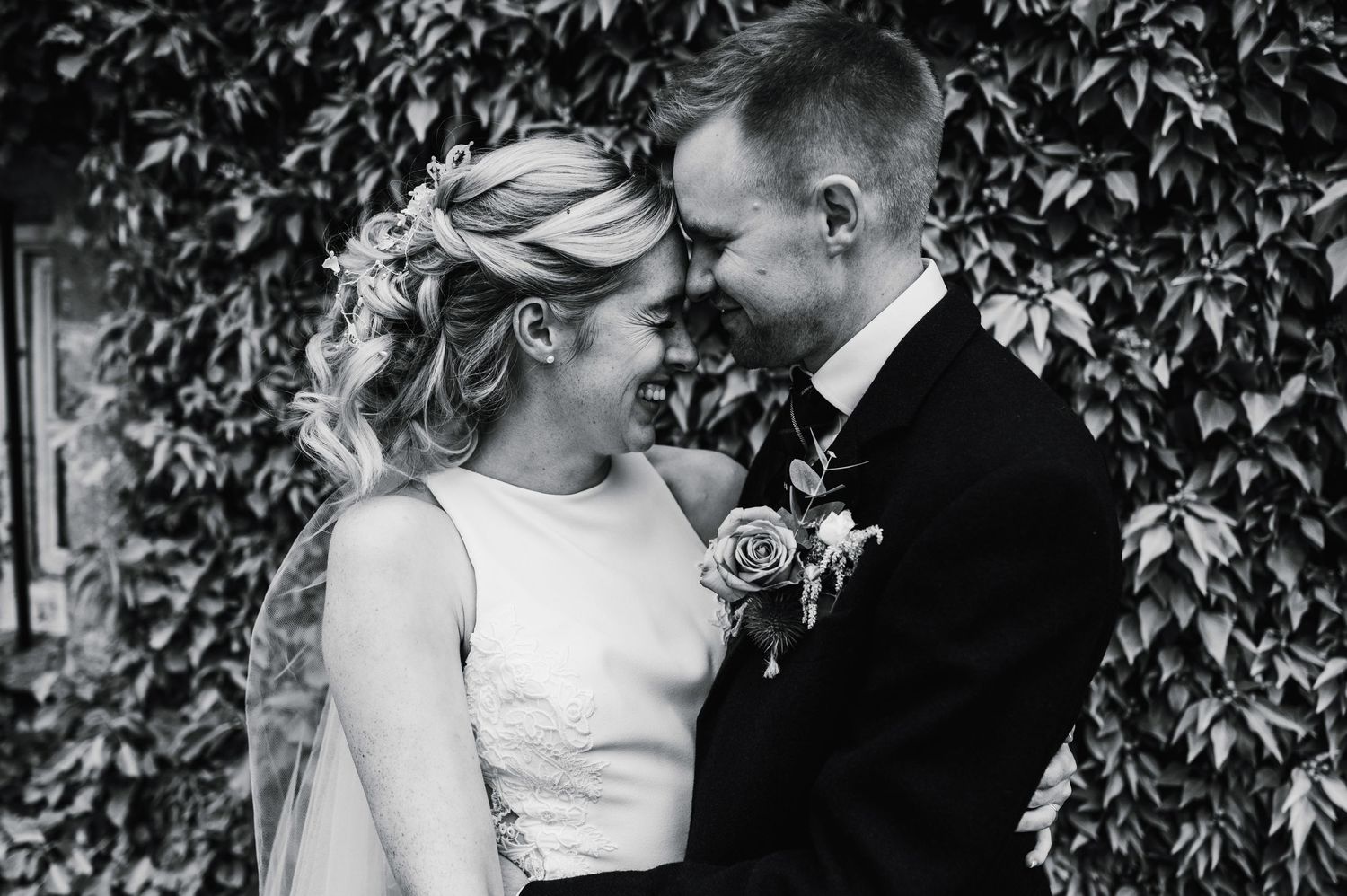 Black and white intimate portrait of a couple in wedding attire sharing a tender moment against a leafy backdrop.