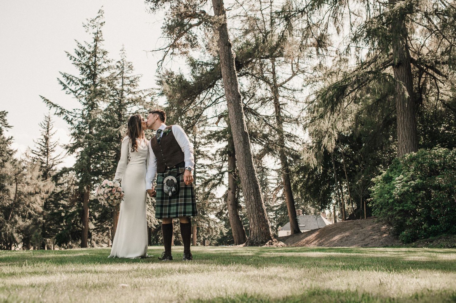 A romantic wedding couple stands among tall evergreen trees wearing Scottish attire in an outdoor forest setting.