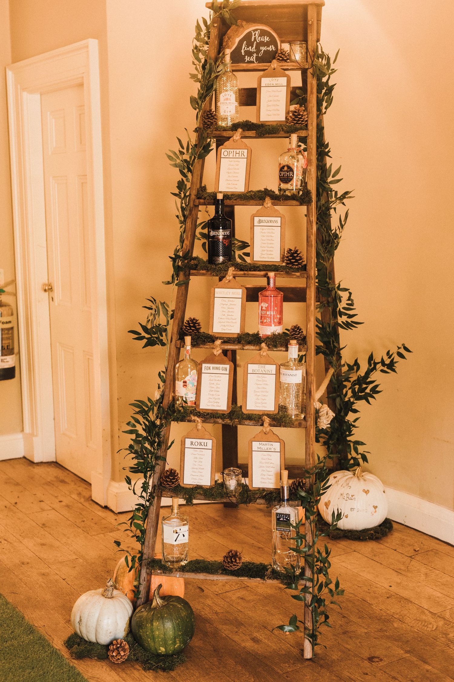 Wooden ladder display featuring wedding seating charts adorned with greenery and white pumpkins.