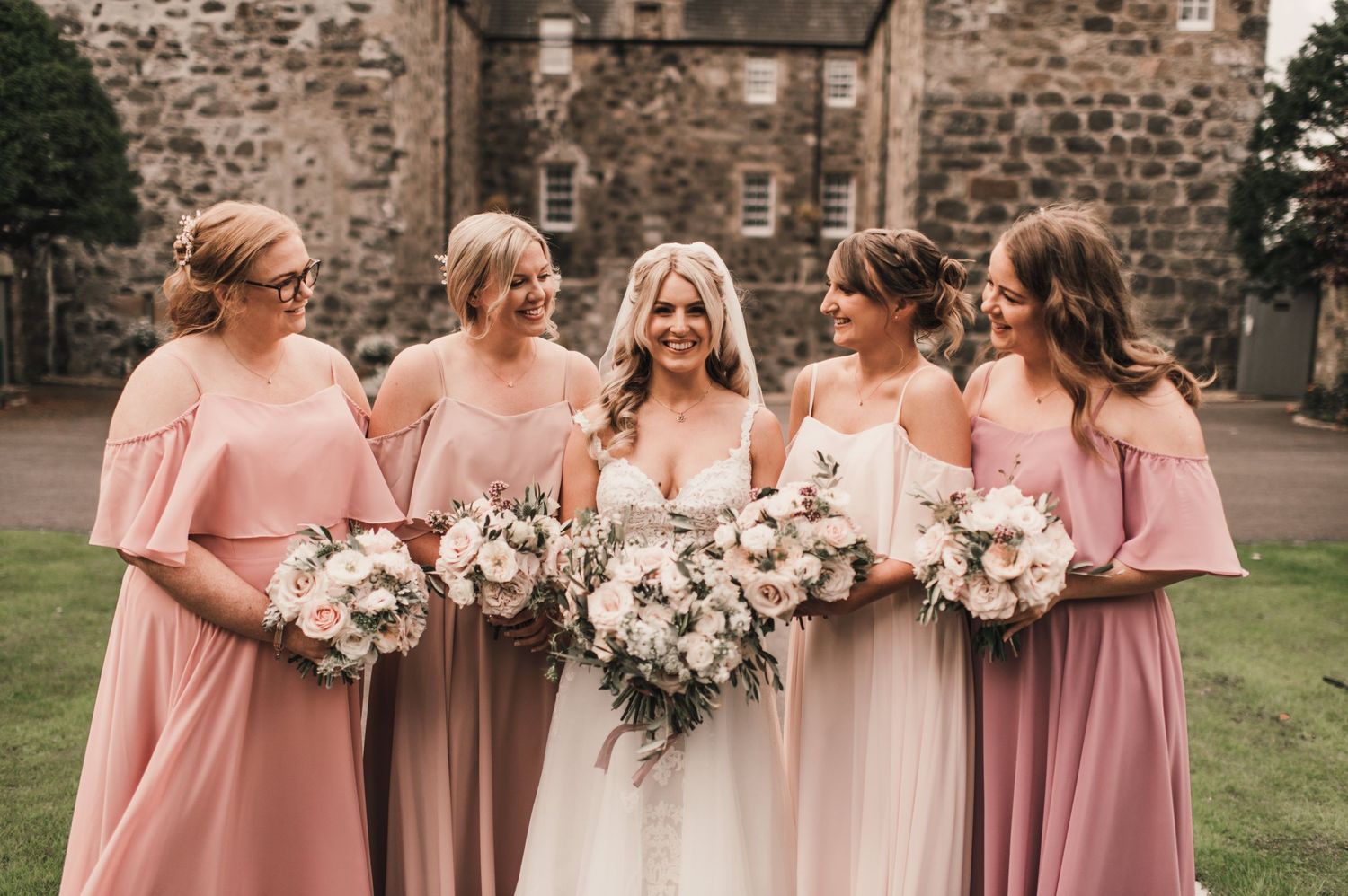 Wedding party wearing pink gowns holding white floral bouquets outside a historic castle.