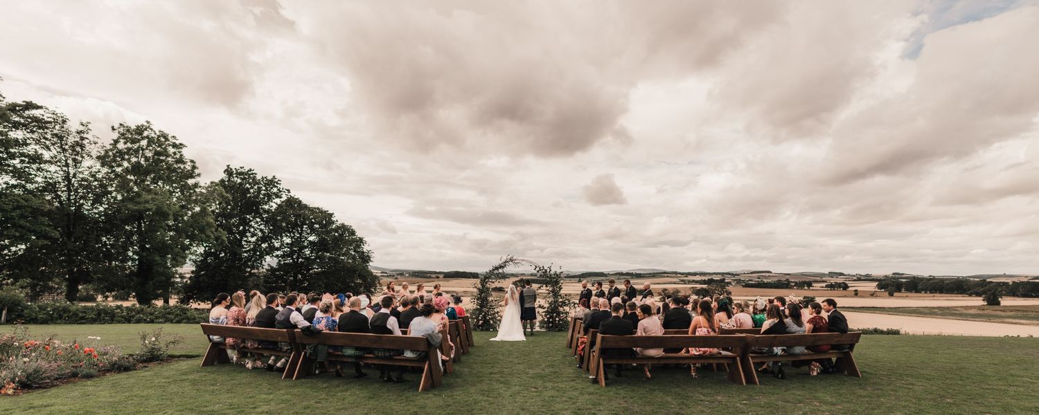 A panoramic view of an outdoor wedding ceremony with guests seated on both sides under cloudy skies.