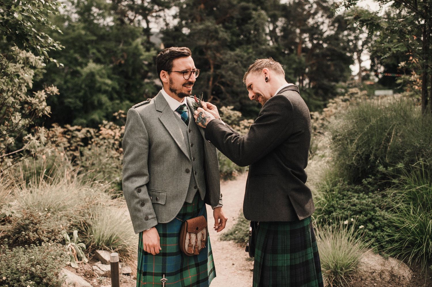 Two people in traditional Scottish wedding attire helping with formal wear adjustments outdoors.