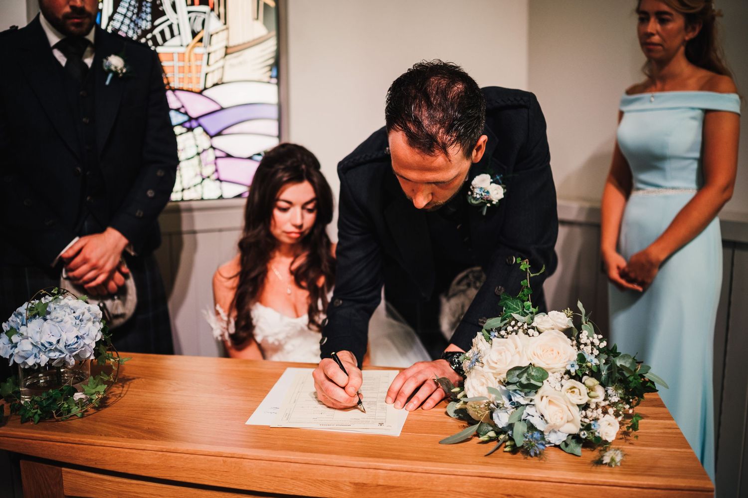 Wedding ceremony signing moment with blue bridesmaids dresses and floral arrangements on wooden table.