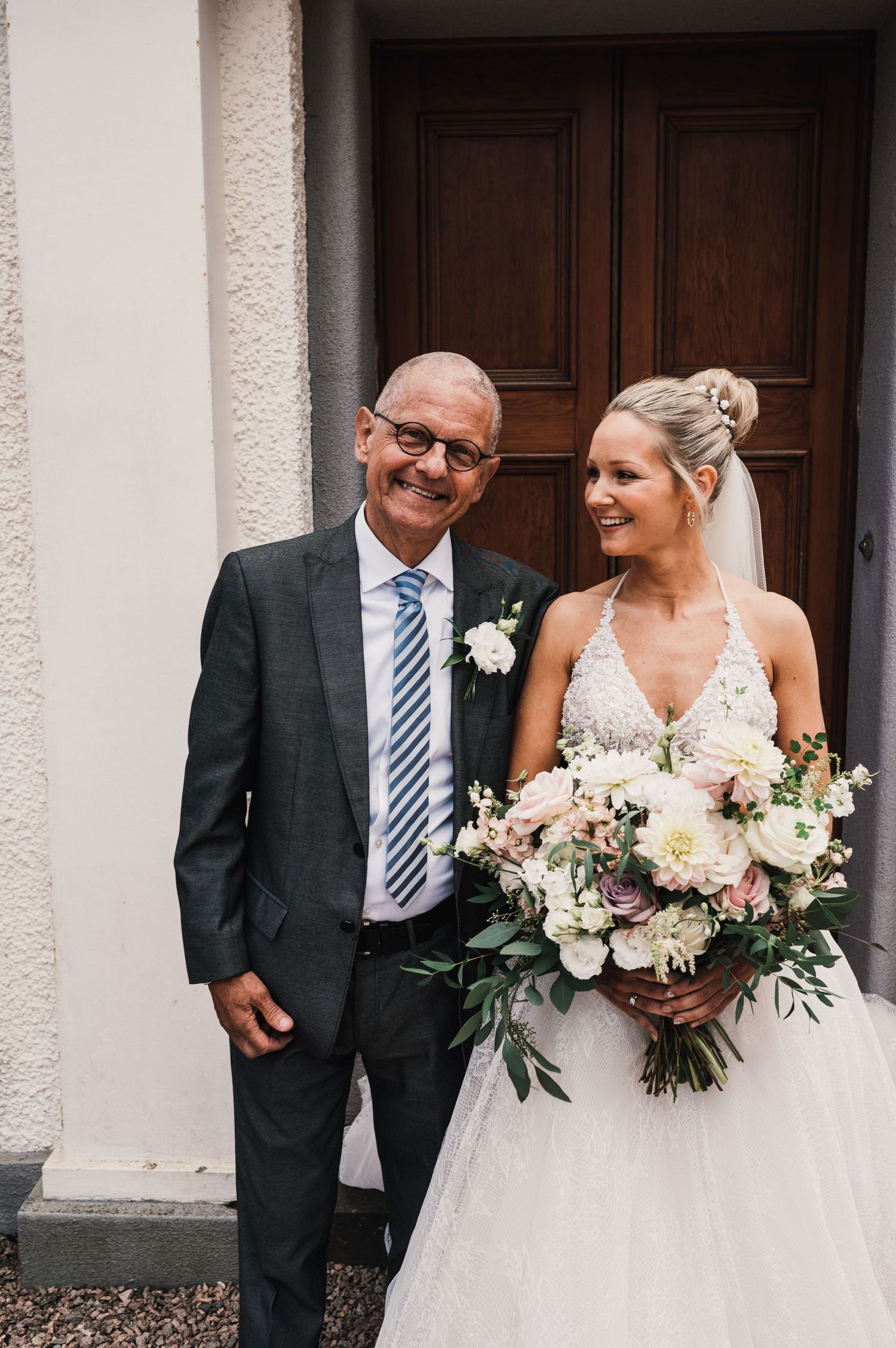 A wedding portrait showing a formal suit and white bridal gown with a lush floral bouquet against wooden doors.