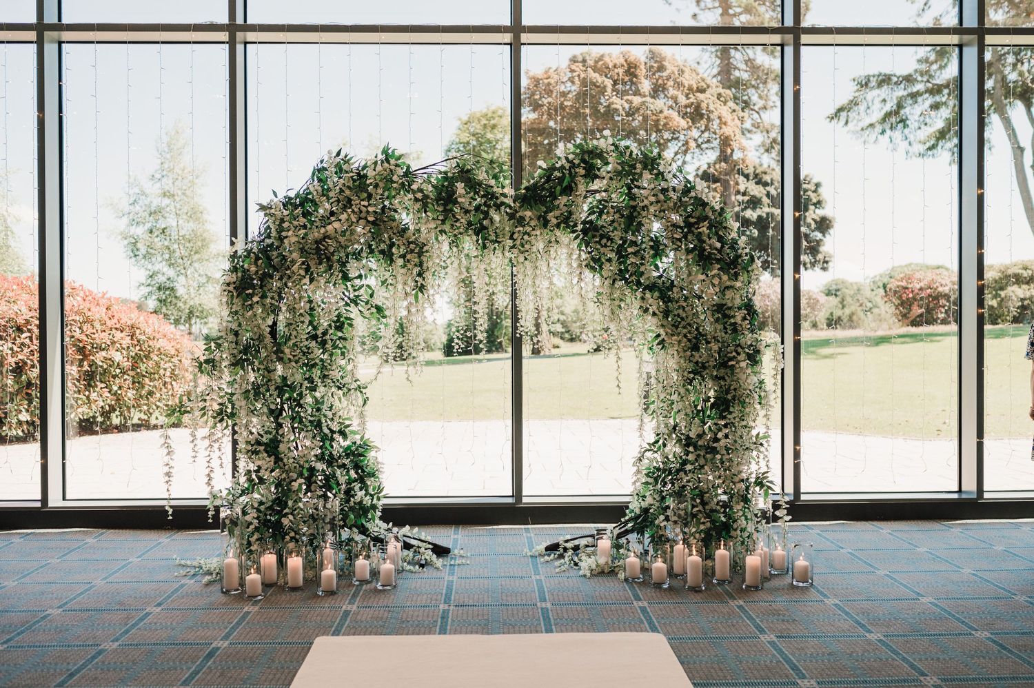 Wedding ceremony arch with cascading greenery and florals against floor-to-ceiling windows.