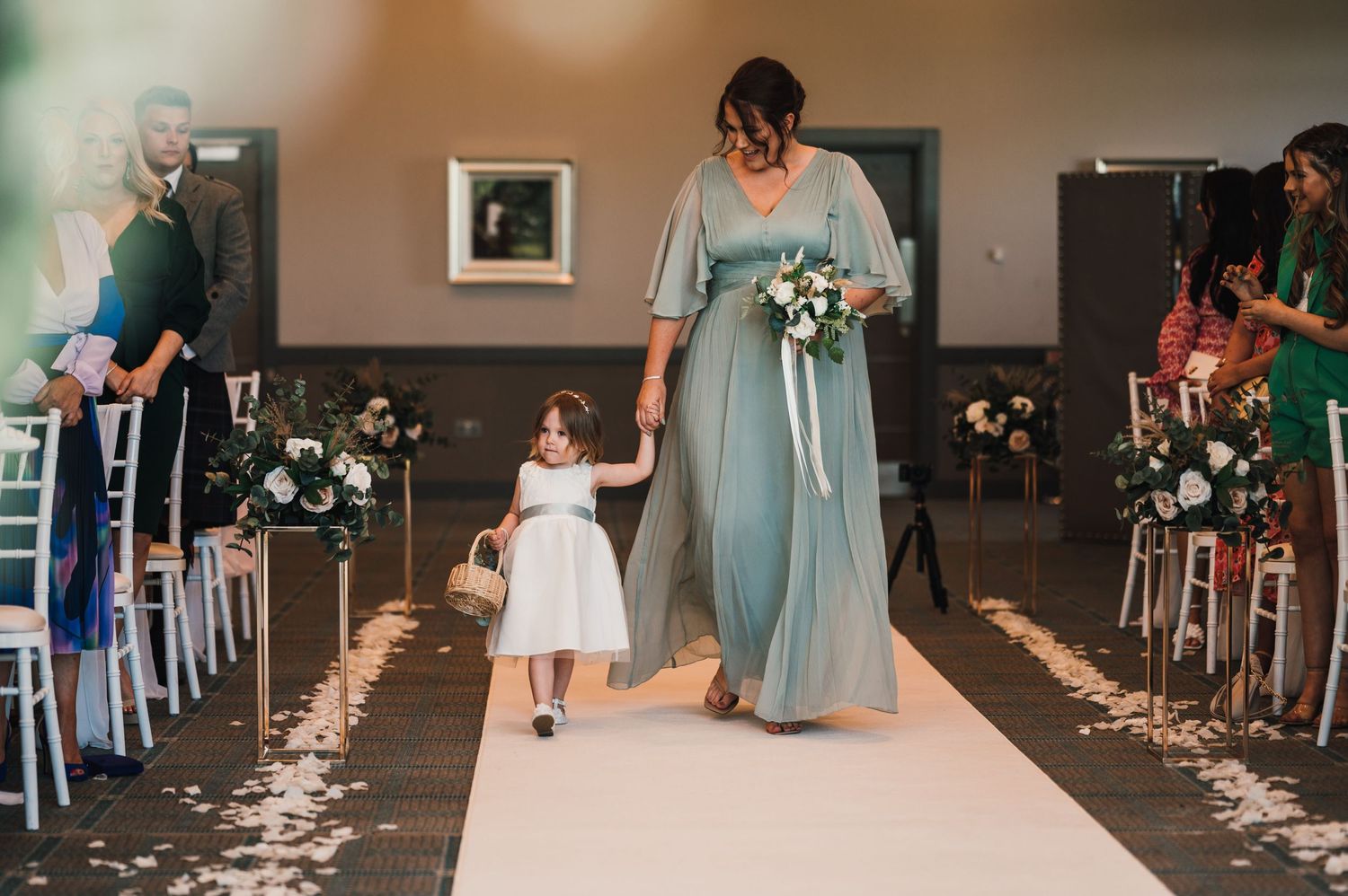 Bridesmaid in blue dress walks with flower girl down petal-strewn aisle.