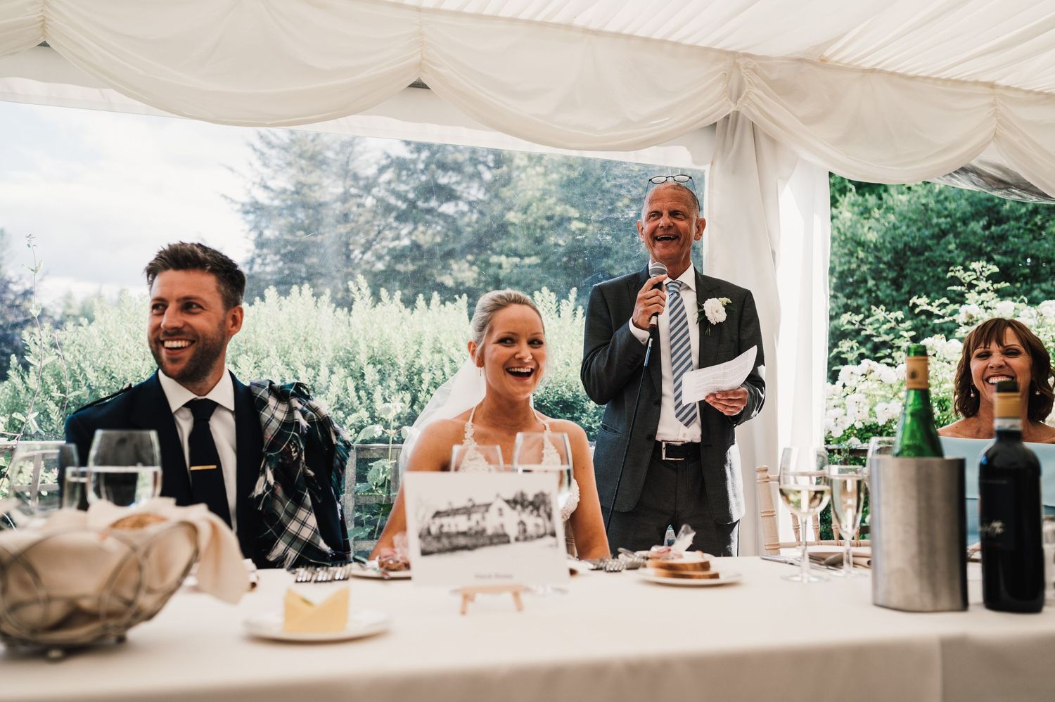 Wedding reception scene with guests enjoying speeches under white tent canopy.