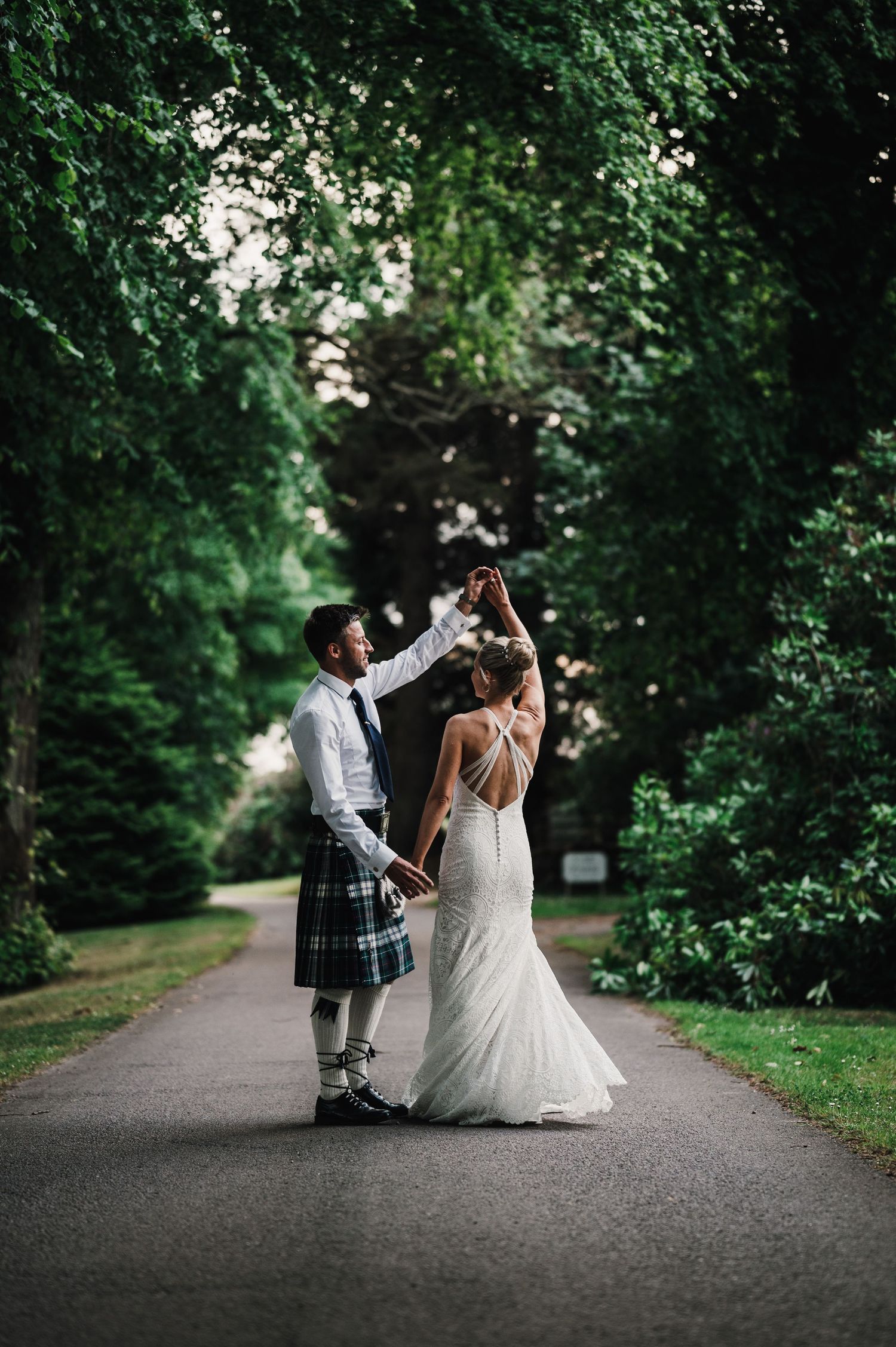 A couple dances on a tree-lined path with the groom wearing a traditional Scottish kilt.