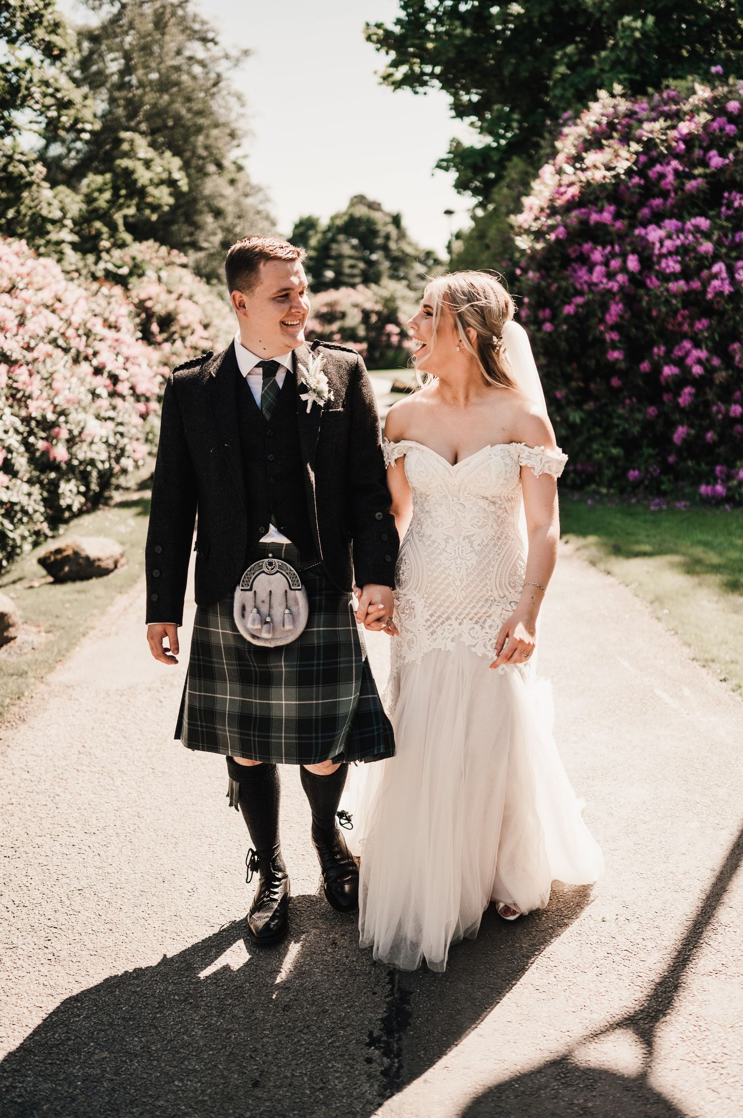 A couple walking along a path with blooming rhododendrons, the groom wearing traditional Scottish attire.