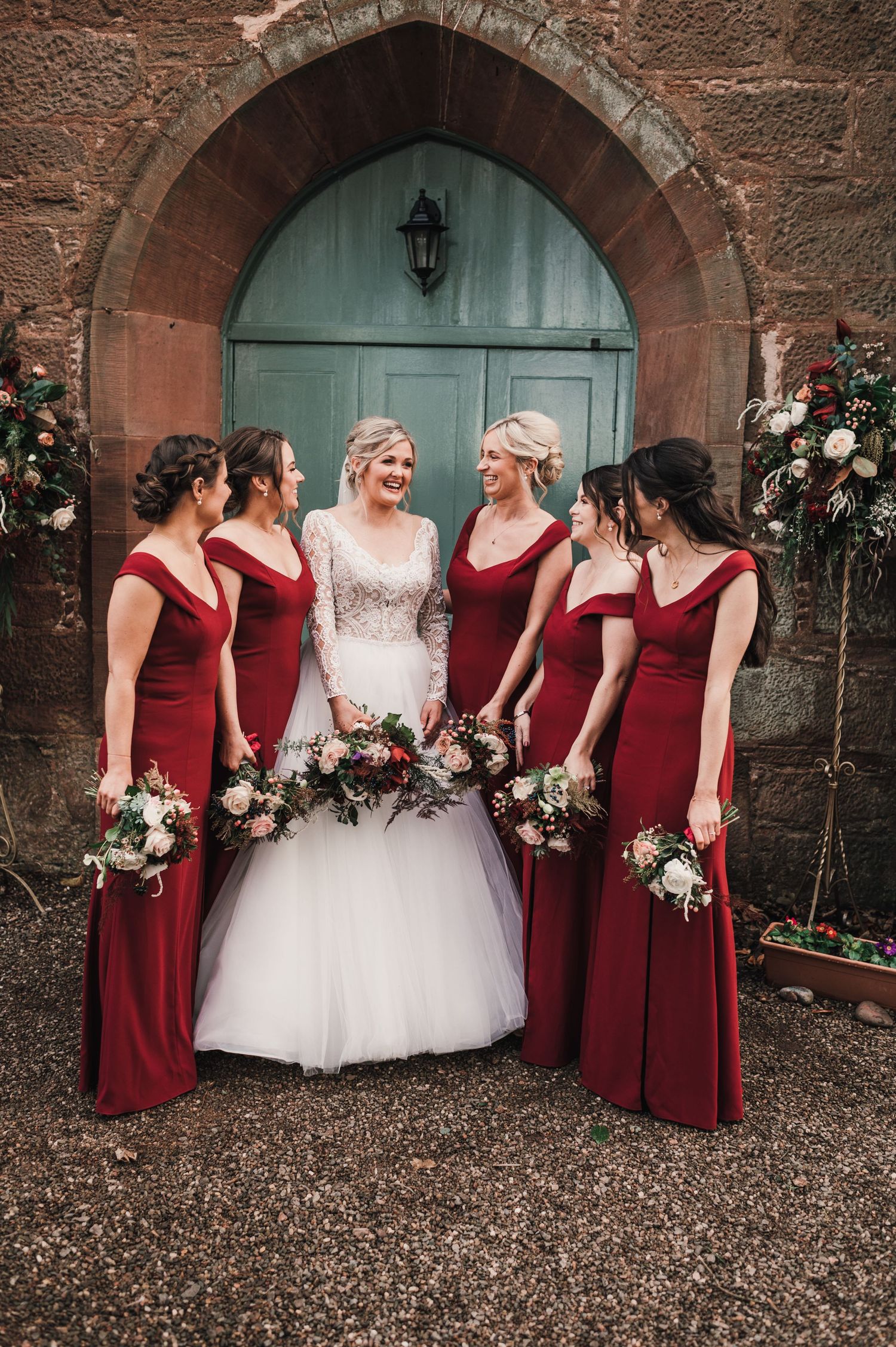 Bride in white gown surrounded by bridesmaids in burgundy dresses at church entrance.