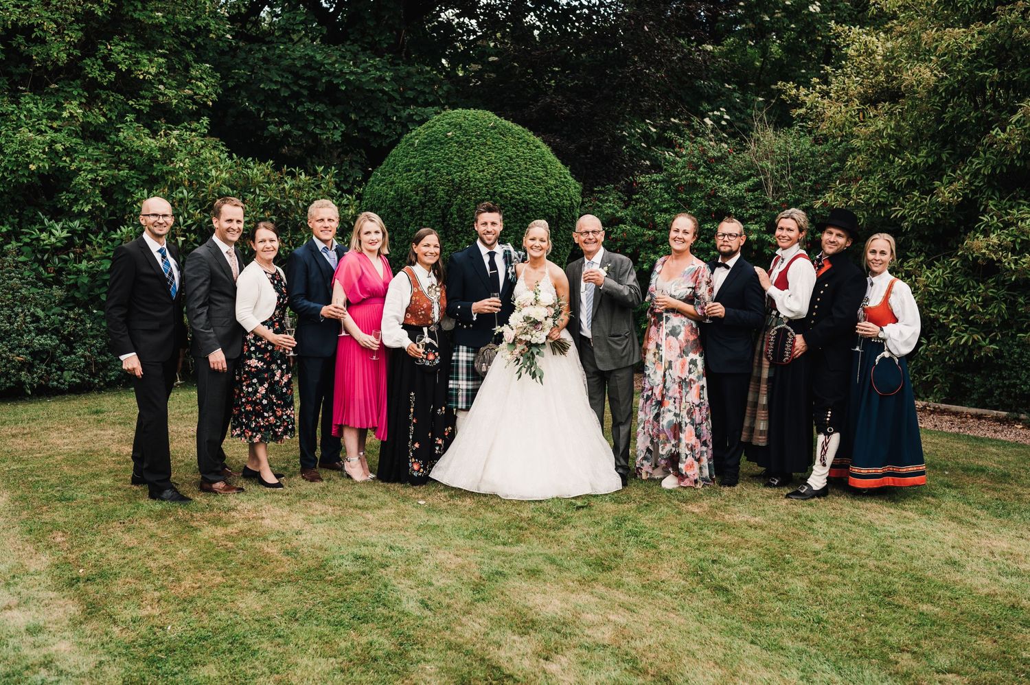 Large wedding party group poses for formal photo on lawn with decorative greenery backdrop.