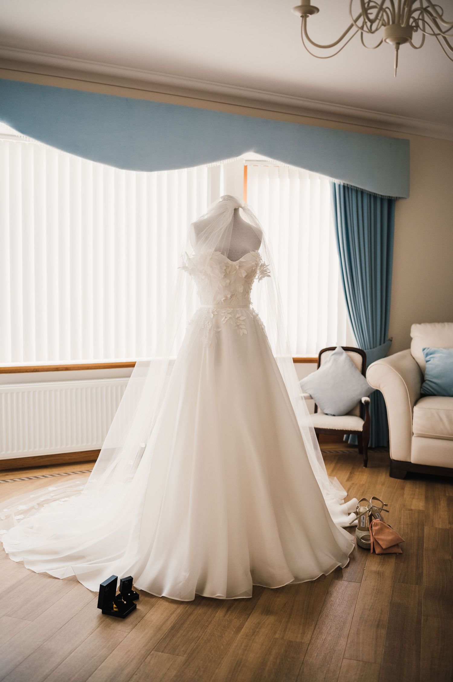 White lace wedding dress hanging in bedroom with blue curtains and hardwood floor.