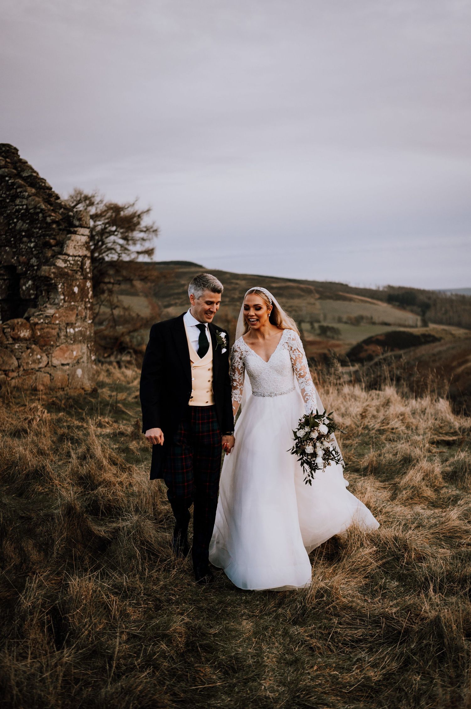 Wedding portrait in countryside setting with rustic stone wall and rolling hills backdrop.