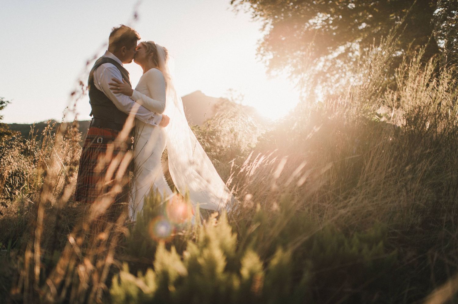 Romantic sunset wedding photo with couple embracing in field of tall grass.