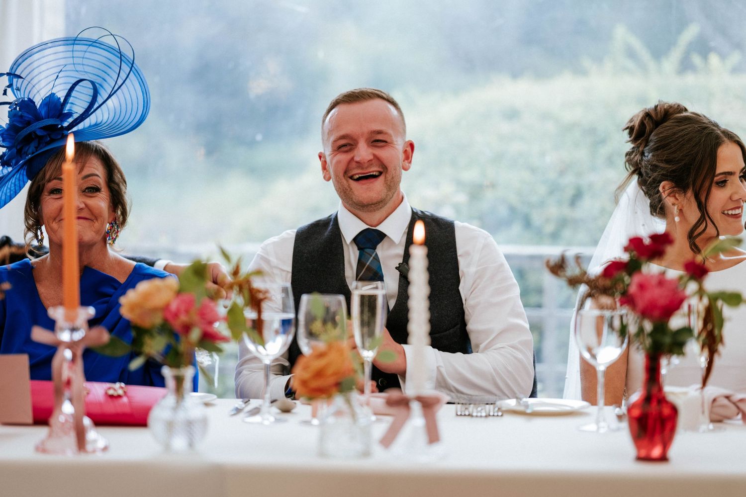Wedding reception guests enjoying dinner and conversation at tables decorated with red floral centerpieces.