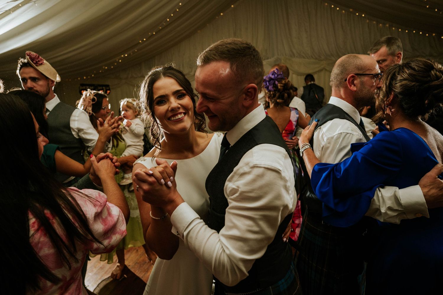 Energetic crowd dancing and celebrating at an indoor wedding reception under draped fabric ceiling.