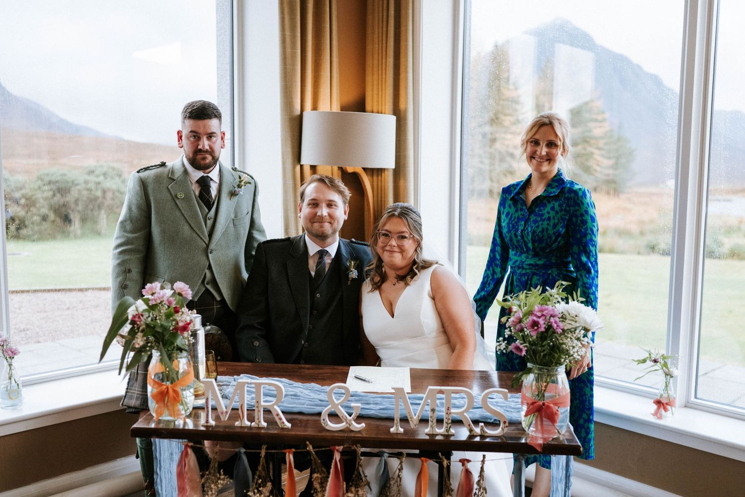 A wedding celebration at a scenic mountain venue with guests gathered around a rustic wooden table with floral centerpieces.