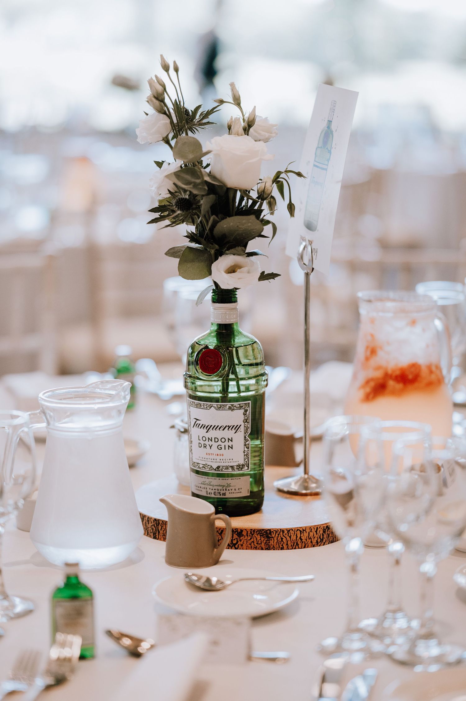 Creative table centerpiece featuring a gin bottle vase with white flowers on rustic wood slice.