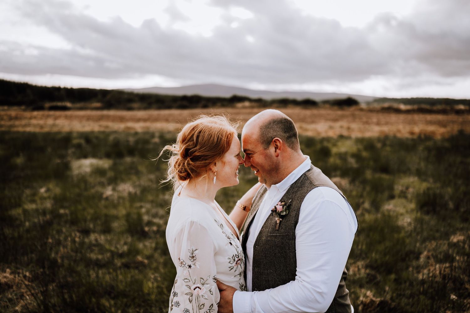 A tender moment between a couple in formal wear standing in a scenic field at dusk.
