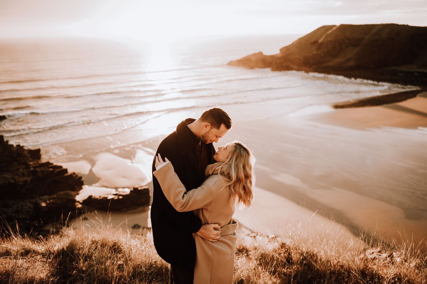 Romantic sunset embrace on coastal clifftop overlooking sandy beach below.