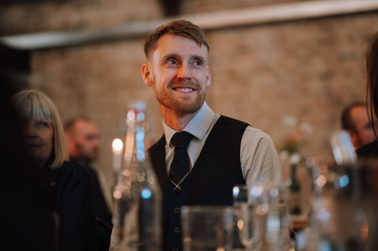 Guest smiles during wedding reception with brick wall and candlelight ambiance.