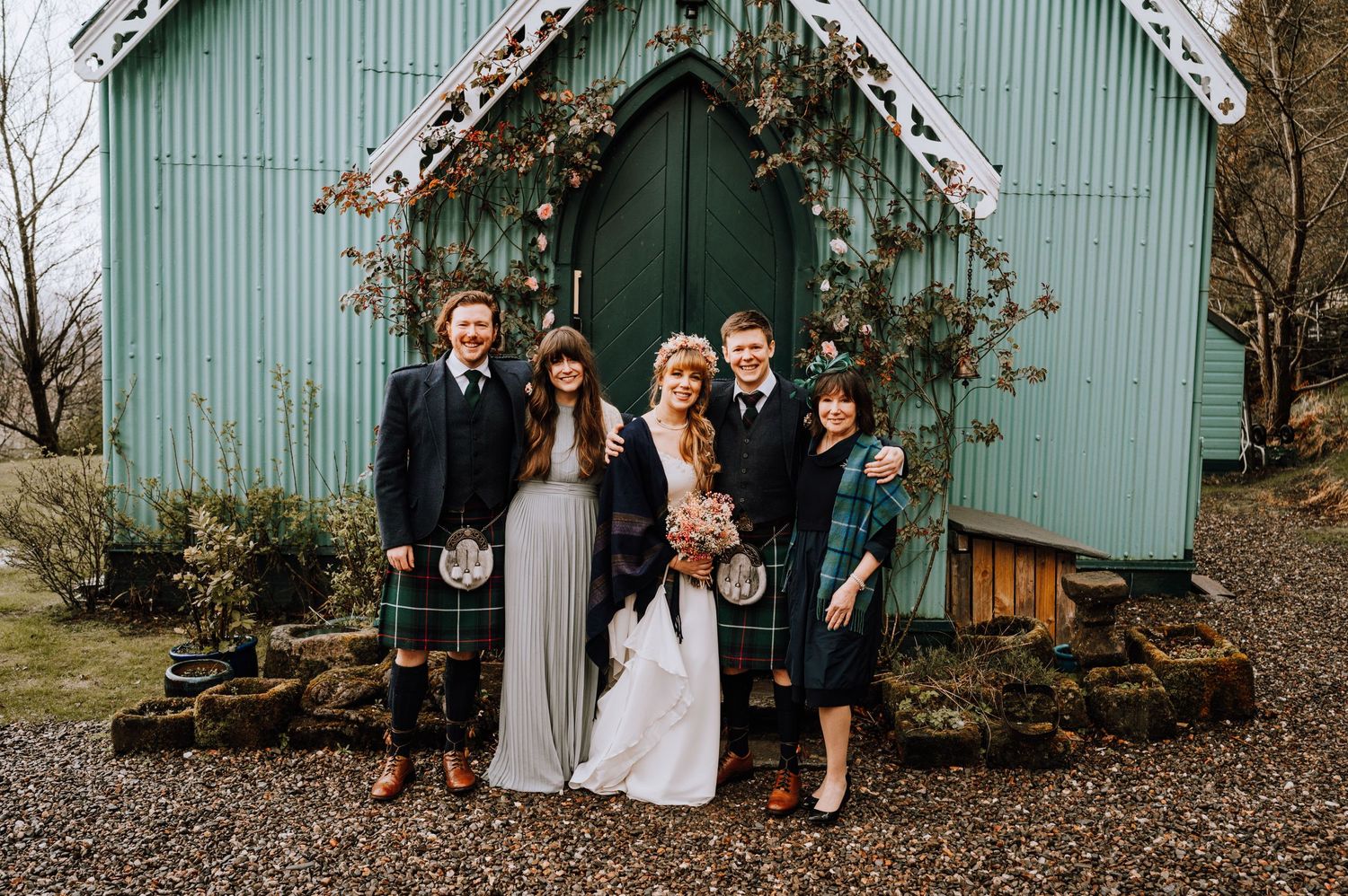 Wedding party poses in front of a teal-colored chapel with a floral archway entrance.