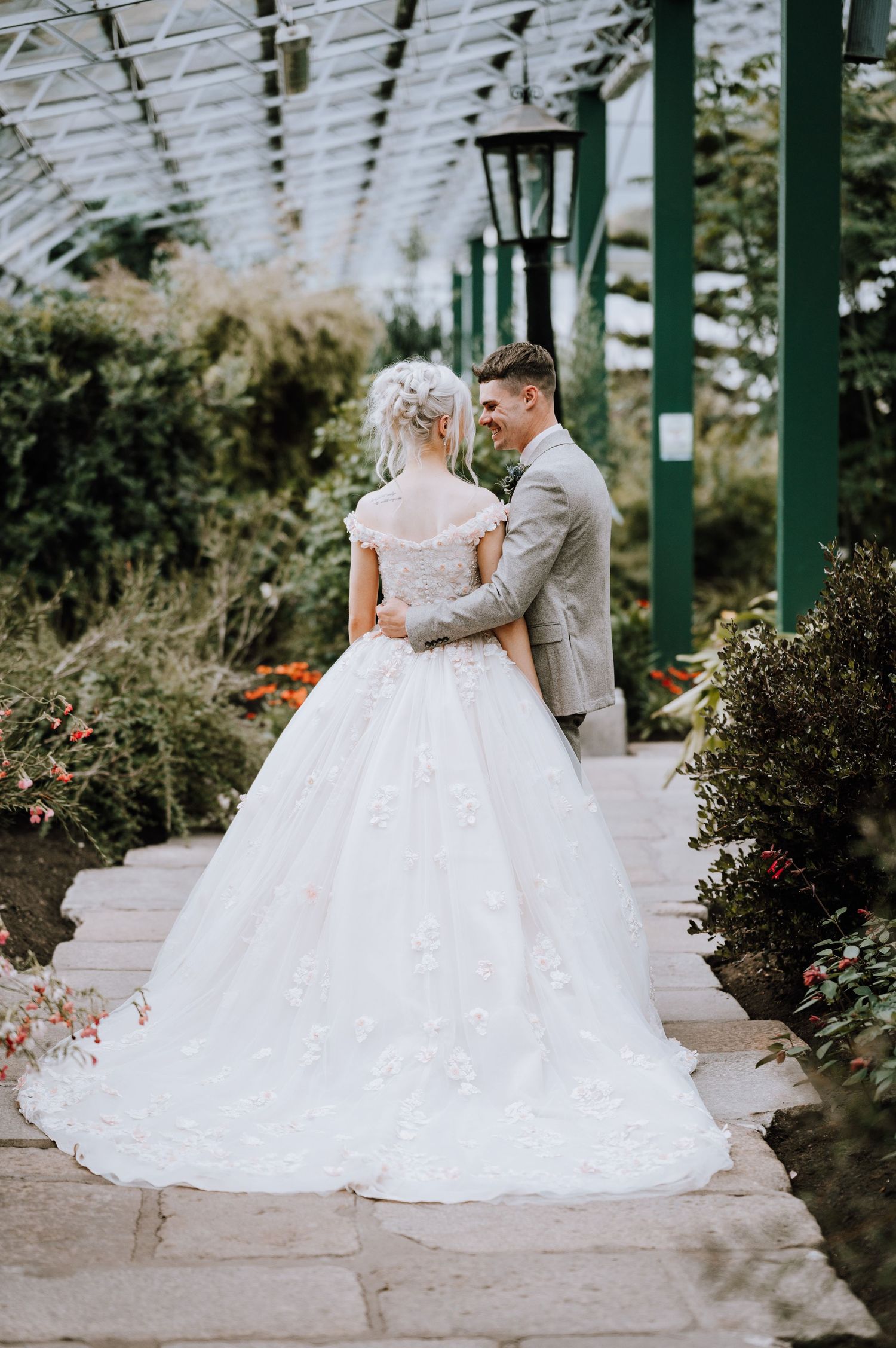 A romantic wedding couple embraces under a glass conservatory roof with trailing greenery.