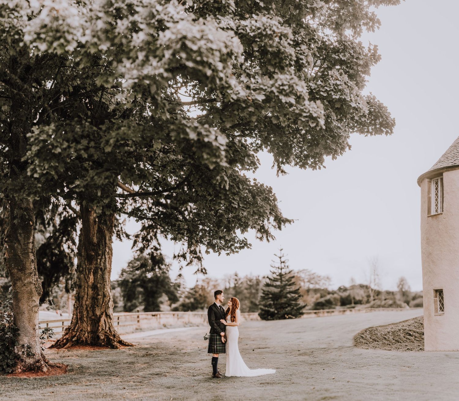 A couple stands beneath a large tree during their outdoor wedding photoshoot.