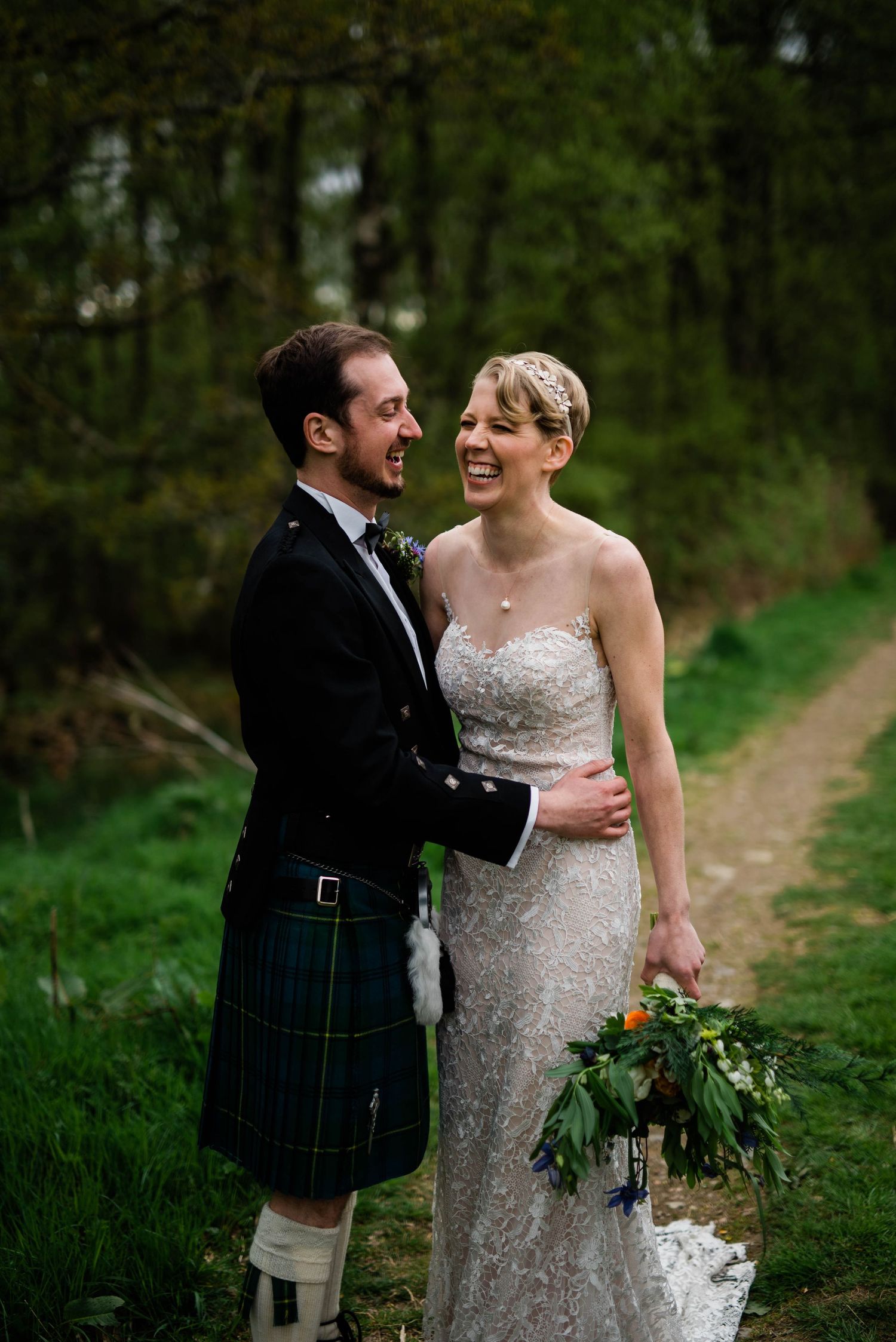 A couple shares a romantic moment on a forest path, wearing wedding attire with Scottish elements.