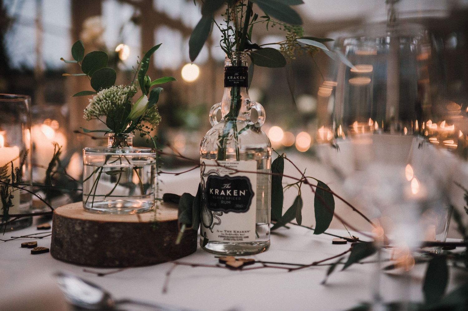 Rustic wedding centerpiece with glass bottles and wooden accents illuminated by string lights.
