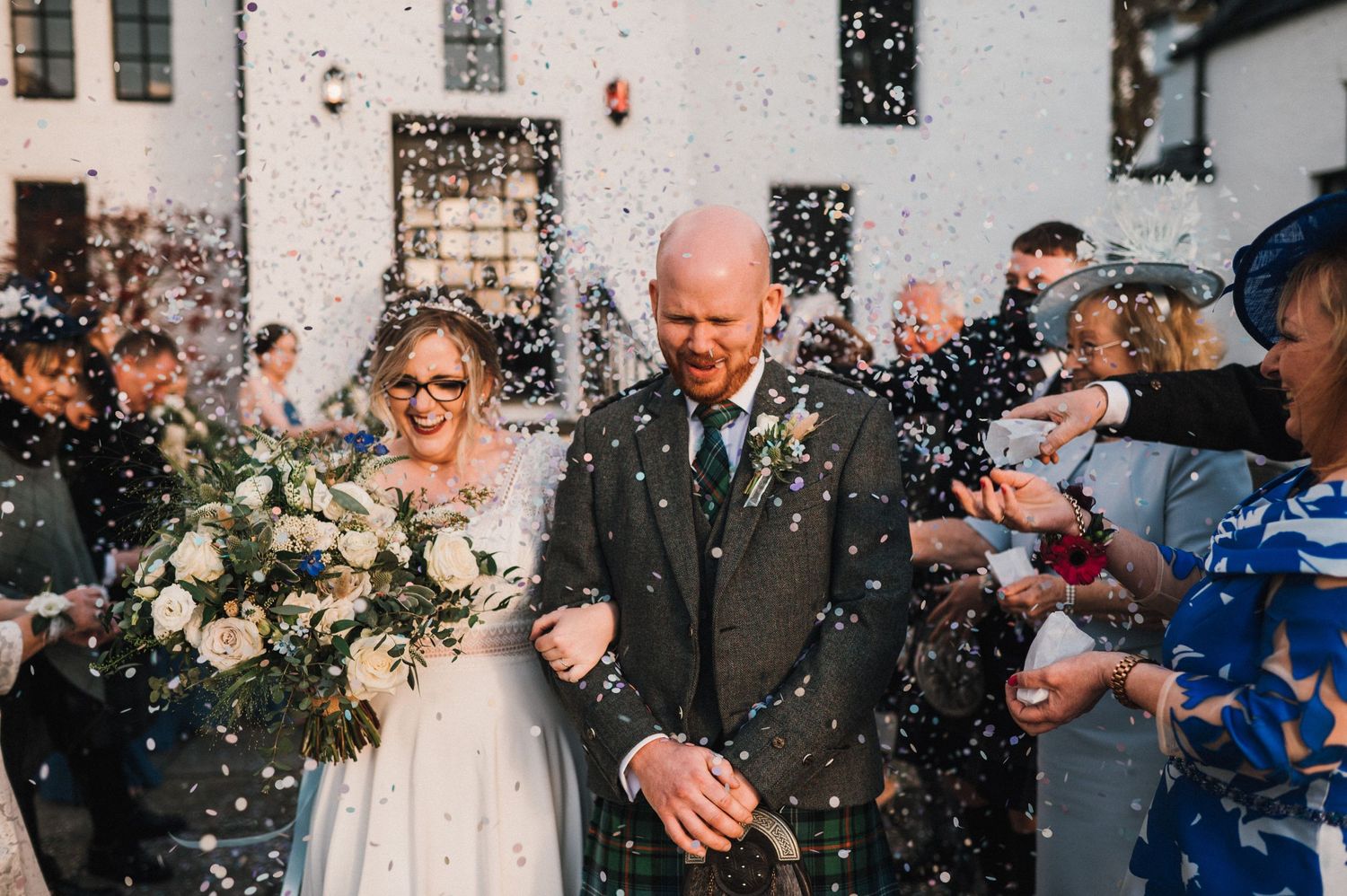 Wedding couple walks through a shower of confetti outside a white building as guests celebrate.